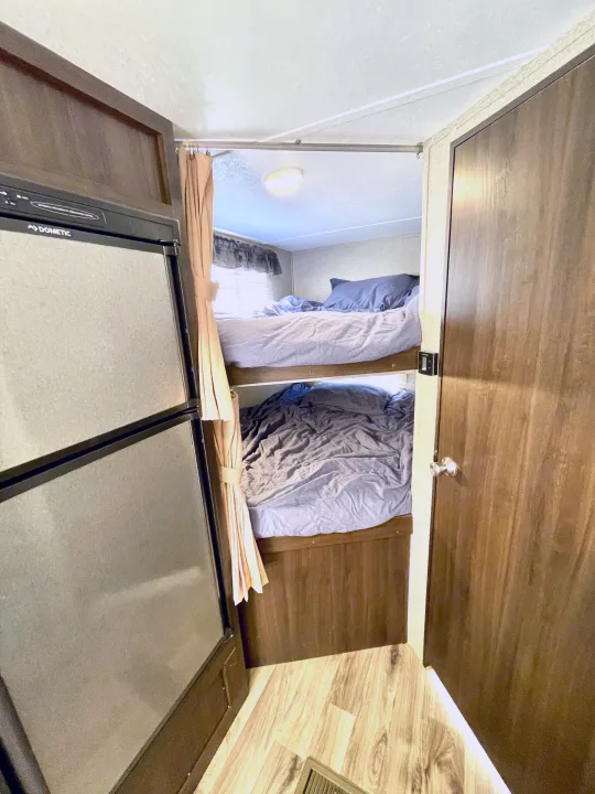RV rental interior: bunk beds with bedding next to a Dometic fridge. A doorway is on the right, offering comfy travel stays.. Keystone RV Springdale Eco-Lite 2018