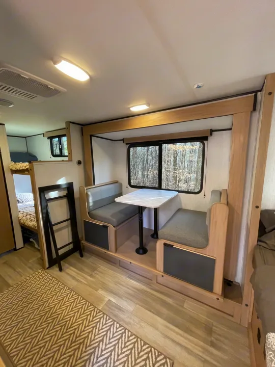 Inside an RV rental: a modern dinette with light wood and gray upholstery, a forest view window, and glimpse of bunk beds.. Cruiser RV Hitch 2022