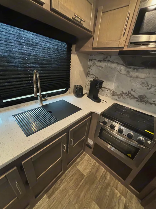 An RV rental kitchen with a sink, stove, microwave, toaster, and coffee maker on a speckled white countertop.. Crossroads RV Sunset Trail Super Lite 2023