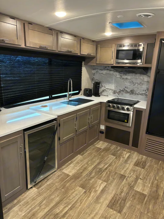 Modern RV rental kitchen with light wood-look cabinets, white countertops, stainless appliances, and a beverage cooler.. Crossroads RV Sunset Trail Super Lite 2023