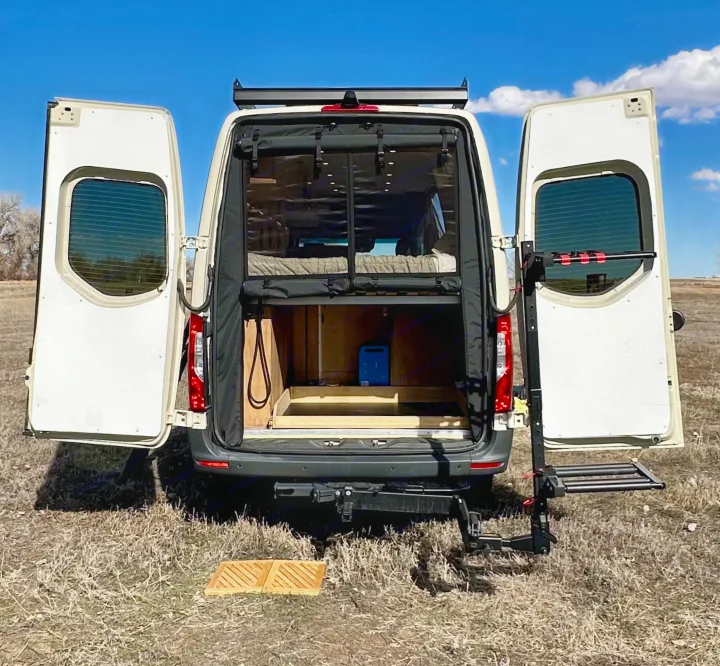 An RV rental camper van with open rear doors reveals an interior bed & storage, parked in a field under blue sky.. Mercedes-Benz Sprinter High Roof Medium wheelbase 2021