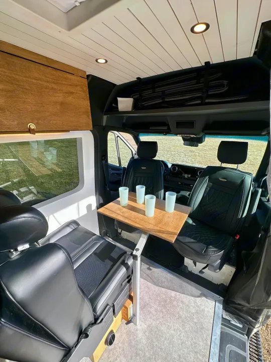 This RV rental shows a modern camper van interior with dining table, comfy seats, and overhead storage.. Mercedes-Benz Sprinter High Roof Medium wheelbase 2021