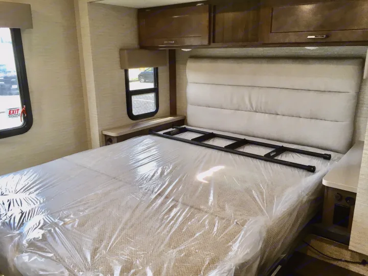 Inside an RV rental bedroom with a new, plastic-wrapped mattress, padded headboard, overhead cabinets, and side windows.. Winnebago Minnie Winnie 2024