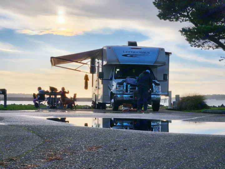 RV rental motorhome parked by water at sunset. People relax under awning, reflections in foreground puddles.. Coachman Leprechaun 2017
