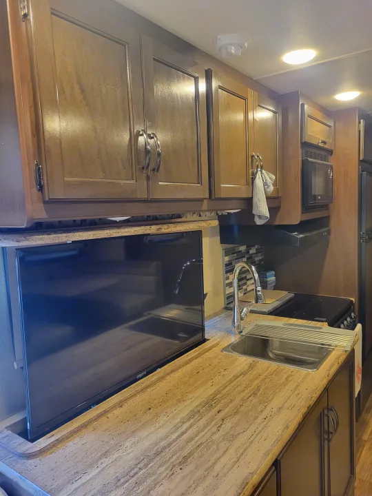 Efficient RV rental kitchen featuring wood cabinets, pop-up TV, sink, and cooktop for comfortable travel.. Thor A.C.E. 2018