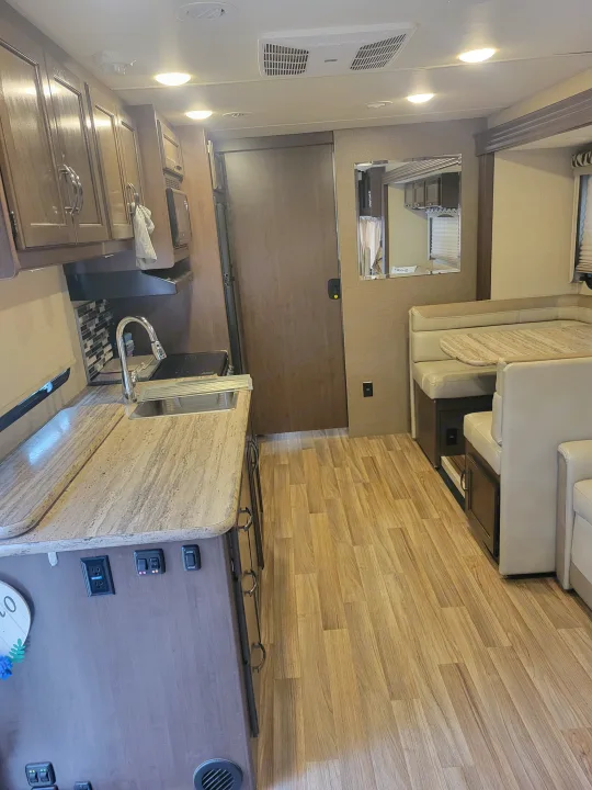 Bright RV rental interior featuring a compact kitchen with sink, cozy booth dinette, and laminate wood flooring.. Thor A.C.E. 2018