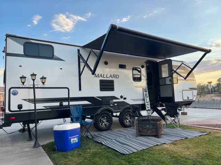 White RV rental deployed with an illuminated awning, outdoor mat, cooler, and fire pit on green grass.. Heartland RVs Mallard 2022