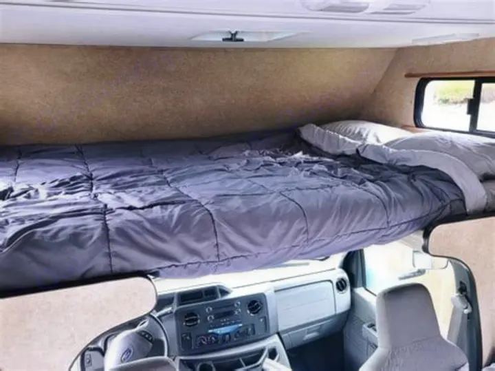 RV rental cab-over bunk: a made-up bed with grey comforter & pillows, above the vehicle's dashboard.. Thor Majestic 2018