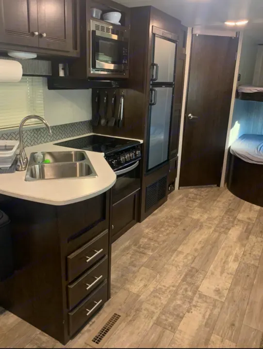 Inside a stylish RV rental: a compact kitchen with dark cabinets, sink, stove, microwave, and fridge, leading to bunk beds.. Heartland Pioneer 2020
