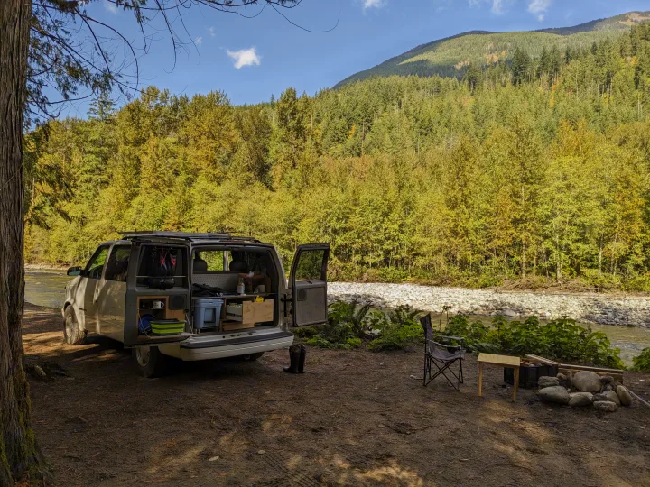 Enjoy a scenic outdoor adventure! This custom camper van (similar to an RV rental) is parked by a river at a forest campsite with mountains.. Chevrolet Open Road 2002