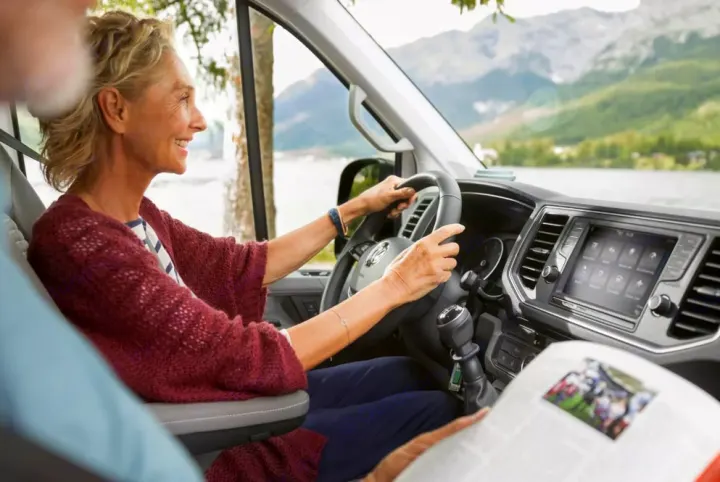 Smiling woman at the wheel of an RV rental, admiring a scenic lake and mountains. A passenger reads nearby.. Volkswagen Grand California 600 boîte automatique 2024