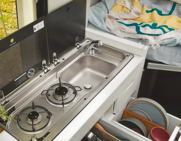 An RV rental's compact kitchen unit: stainless steel sink, 2-burner stove, and open drawers revealing neatly stored dishes.. Volkswagen Grand California 600 boîte automatique 2024