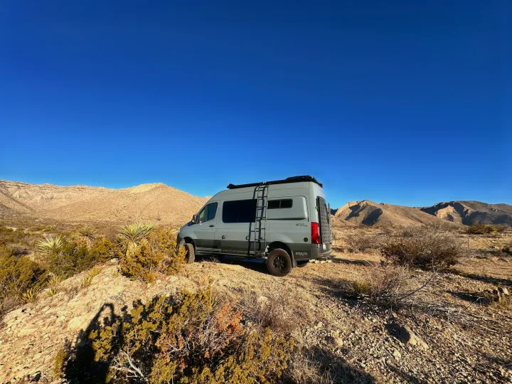 This grey Storyteller Overland 4x4 adventure van, ideal for an RV rental, is parked on a desert hill with arid mountains.. Storyteller Overland Storyteller Overland Class B 2023