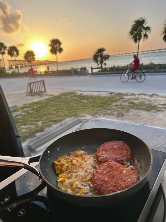 RV rental cooking at sunset: sizzling burgers & eggs by the beach, with a bridge and passing cyclists.. Storyteller Overland Storyteller Overland Class B 2023