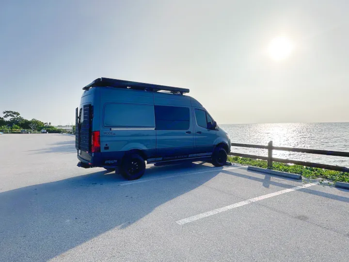 This Storyteller Overland camper van by the water, ideal for an rv rental adventure. Sun glints on the waves.. Storyteller Overland Storyteller Overland Class B 2023