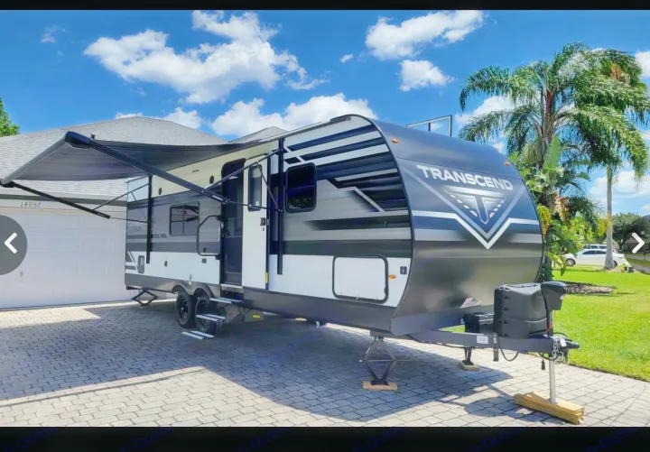 A sleek "TRANSCEND" travel trailer, ready for your next RV rental adventure, parked on a residential driveway with its awning extended.. Grand Design Other 2022