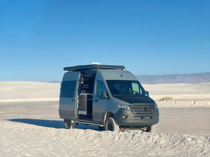A gray Mercedes Sprinter van, an RV rental, is parked on white sand dunes with its side door open, revealing the interior.. Storyteller Overland Storyteller Overland Class B 2023