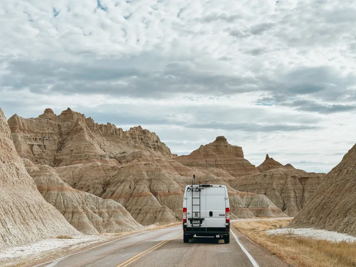 RV rental camper van drives through Badlands National Park, surrounded by unique eroded landscapes under a cloudy sky.. RAM ProMaster 2500 2022