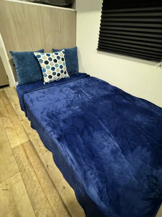 Plush blue bed in an RV rental sleeping area, complete with decorative pillow, wood paneling, and privacy blinds.. Coleman Lantern LT 17B 2024