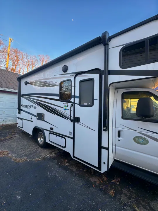 A white Sunseeker Class C RV rental is parked on asphalt. Side view shows an extended awning and the moon overhead.. Forest River Sunseeker LE 2022