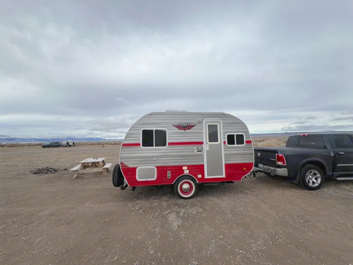 Retro RV rental hitched to a pickup truck, parked by a picnic table in a vast, cloudy landscape with distant mountains.. Riverside Travel Trailer 157 Retro 2018