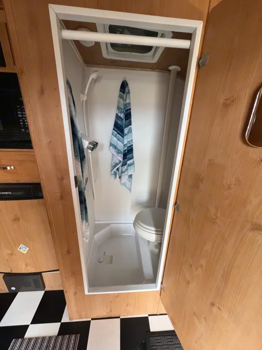 View inside a compact RV rental bathroom, featuring a shower, toilet, ceiling vent, and checkered floor.. Riverside Travel Trailer 157 Retro 2018