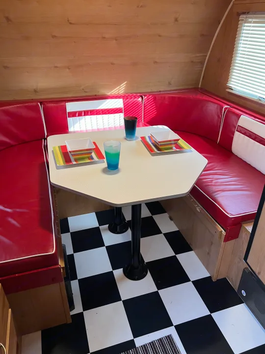 An RV rental interior with a retro red diner booth, octagonal table set with colorful dishes, and a black and white checkered floor.. Riverside Travel Trailer 157 Retro 2018