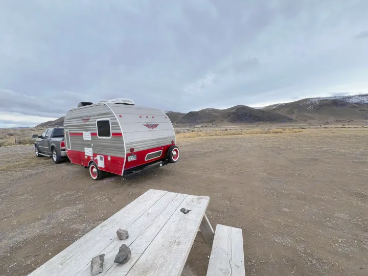 This retro red & gray rv rental travel trailer, with a truck, is parked on a dirt lot near a picnic table, under cloudy skies.. Riverside Travel Trailer 157 Retro 2018
