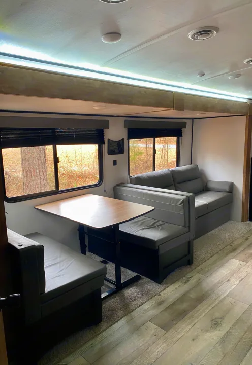 Inside an RV rental: a modern living space with a dining booth, sofa, and forest views from large windows.. Keystone RV Springdale 2022