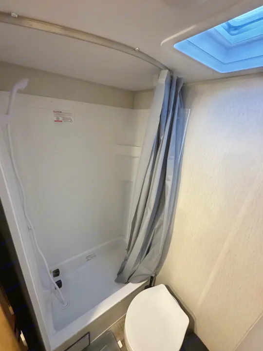 RV rental bathroom with a white shower stall, curved gray shower curtain, and a toilet. A skylight brightens this compact space.. Forest River Cherokee Wolf Den 16 EV 2024