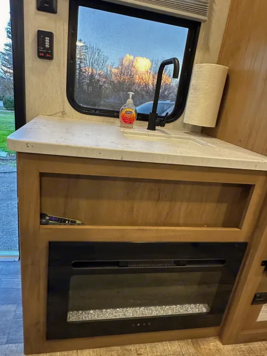RV rental interior: a compact counter with sink, soap, paper towels, window view, control panel, and electric fireplace.. Forest River Cherokee Wolf Den 16 EV 2024