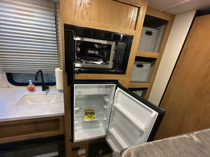 This rv rental features a compact kitchen with a microwave, open fridge, sink, and built-in storage with decorative bins.. Forest River Cherokee Wolf Den 16 EV 2024