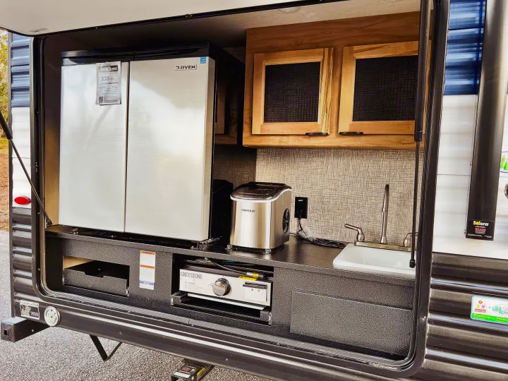 This rv rental outdoor kitchen features a stainless fridge, sink, Greystone pull-out grill, Contoure ice maker, and cabinets.. Forest River Cherokee Grey Wolf 2024