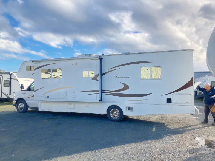 White Adventurer RV motorhome (rv rental) with extended slide-out on gravel, under a cloudy sky. Man inspects it.. Adventurer Adventurer Motorhome 2012