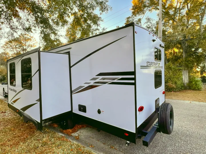 Exterior view of a white Starcraft Autumn Ridge RV rental with its slide-out extended, parked by a road with trees overhead.. Starcraft Autumn Ridge 2024