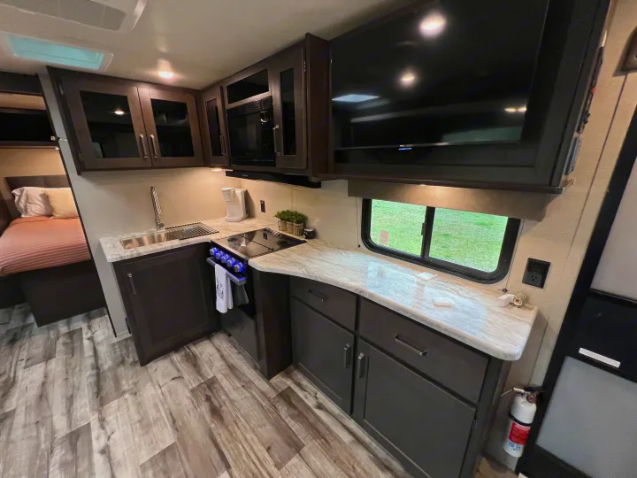 Spacious RV rental interior featuring a full kitchen with microwave, stove, sink, and cabinets, plus a cozy bed area and TV.. Grand Design Transcend Xplor 2023