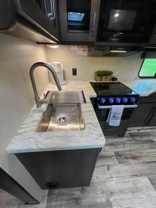 Modern RV rental kitchen setup with a stainless steel sink, blue-lit stove, microwave, and light-toned countertops.. Grand Design Transcend Xplor 2023