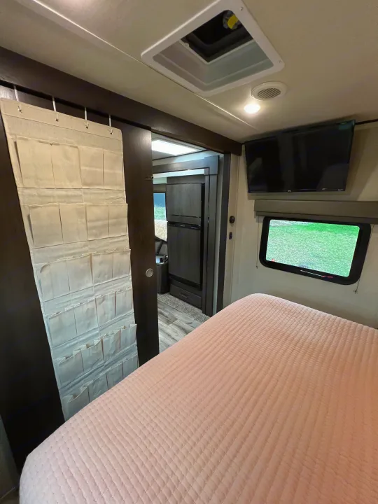 RV rental: Bedroom interior featuring a pink quilted bed, hanging storage organizer, wall-mounted TV, and window.. Grand Design Transcend Xplor 2023