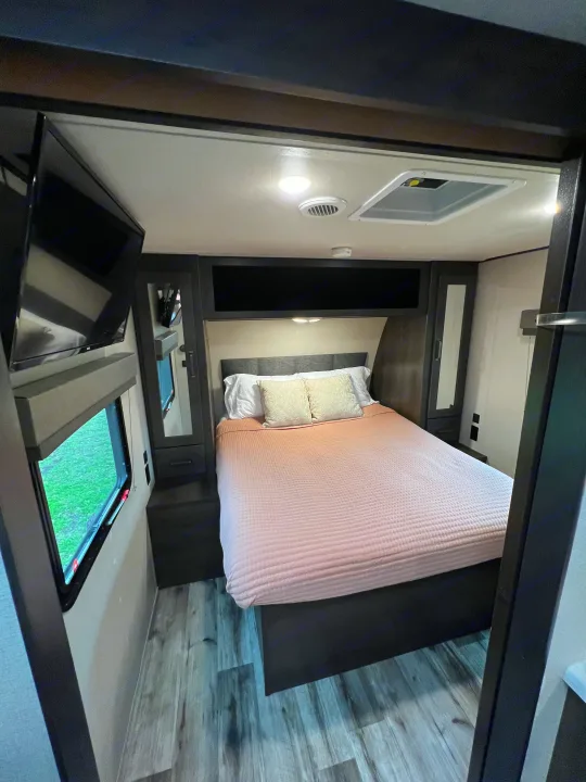 Cozy RV rental bedroom featuring a comfortable bed, wall-mounted TV, window, and built-in storage solutions.. Grand Design Transcend Xplor 2023