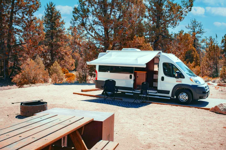 RV rental campervan parked at a scenic campsite with an extended awning, chairs, picnic table, and fire pit setup.. Dodge Promaster 2500 2014