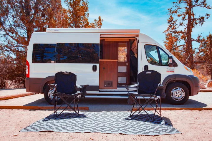 RV rental: A white campervan with its side door open is parked at a campsite, showing two camping chairs and a mat outside.. Dodge Promaster 2500 2014