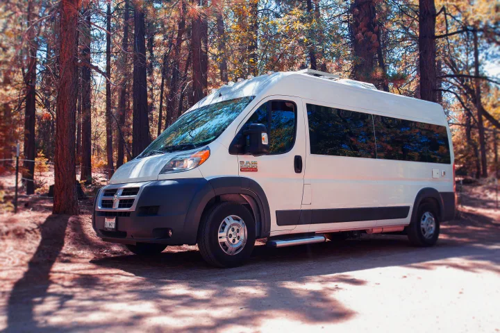 White Ram ProMaster RV rental parked on a forest trail under dappled sunlight, equipped with a roof AC and side step.. Dodge Promaster 2500 2014