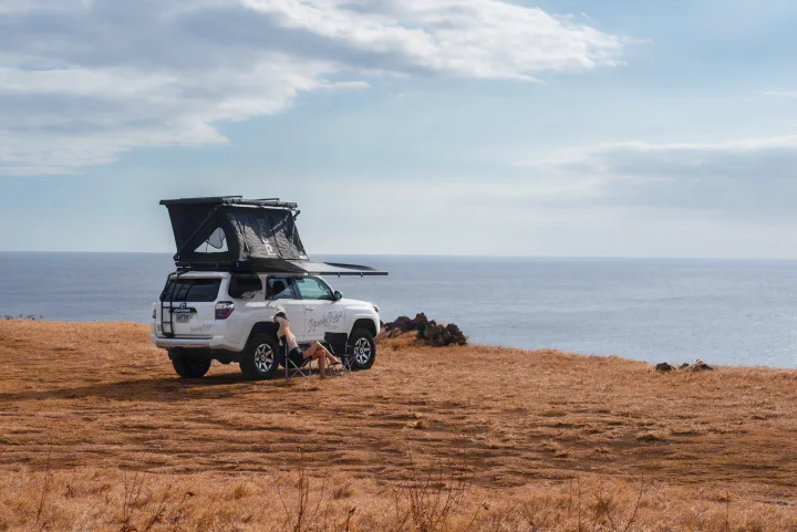 Ocean views from this RV rental! White 4Runner with rooftop tent & awning on a cliff. Person relaxing by the sea.. Custom Toyota 4Runner TRD Off-Road 2023