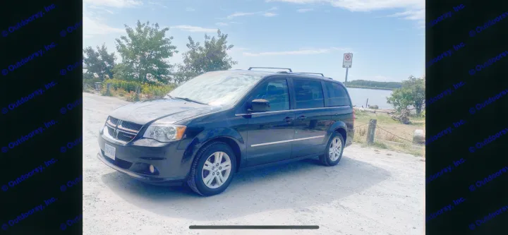 Black minivan parked on a sunny gravel path by the water. A practical RV rental for road trips & outdoor exploration.. Dodge Grand Caravan 2015