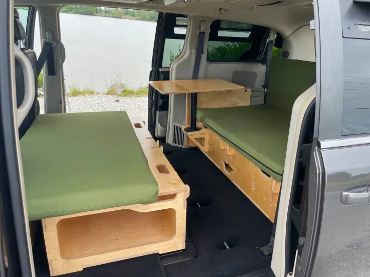 This rv rental features a minivan camper conversion with two green-cushioned beds, a fold-out table, and water views. Perfect for road trips.. Dodge Grand Caravan 2015