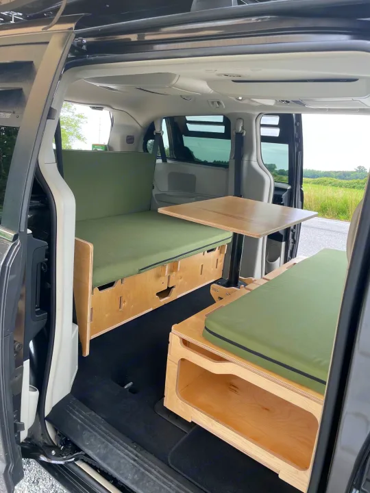 Inside a custom rv rental minivan camper: wooden benches with green cushions, a table, and open door to nature.. Dodge Grand Caravan 2015