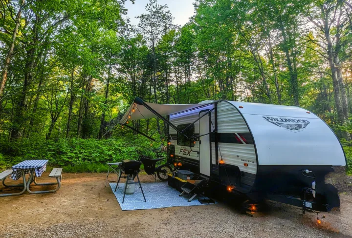RV rental: Cozy camper with lit awning, picnic table, grill, and bike set up on a dirt campsite surrounded by lush forest.. Forest River Wildwood FSX 2021