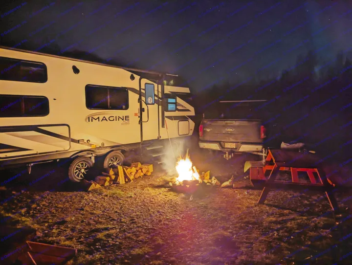 Cozy night camping scene: RV rental & truck parked, warm campfire, stacked wood, picnic table, and starry sky.. Grand Design Imagine XLS 2023