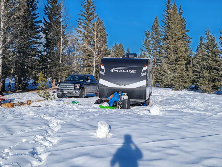 An rv rental (Imagine XLS travel trailer) and a black truck are parked in a snowy winter campground with tall trees.. Grand Design Imagine XLS 2023