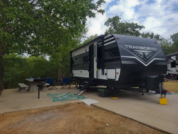 An RV rental setup: a black & white Transcend travel trailer, picnic table, and chairs at a lush campsite.. Grand Design Transcend Xplor 2021